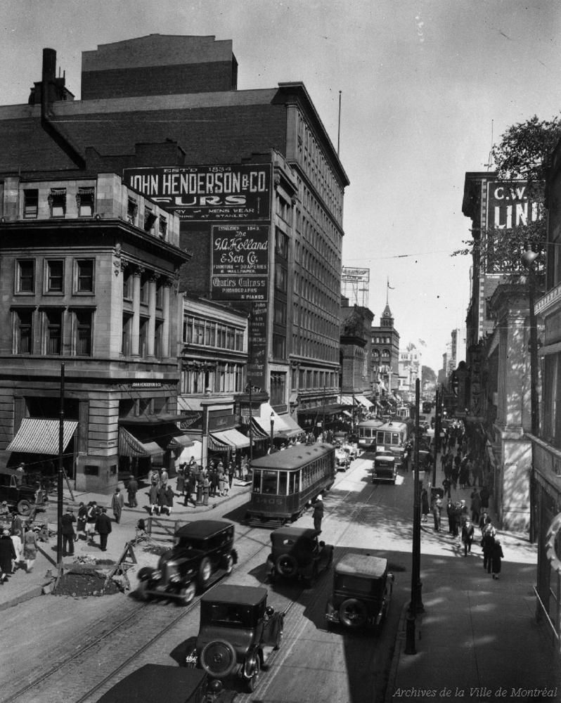 A bustling city street with an eight or nine storey building, likely a department store; lots of pedestrians; and the street full of 1930s cars and tramcars